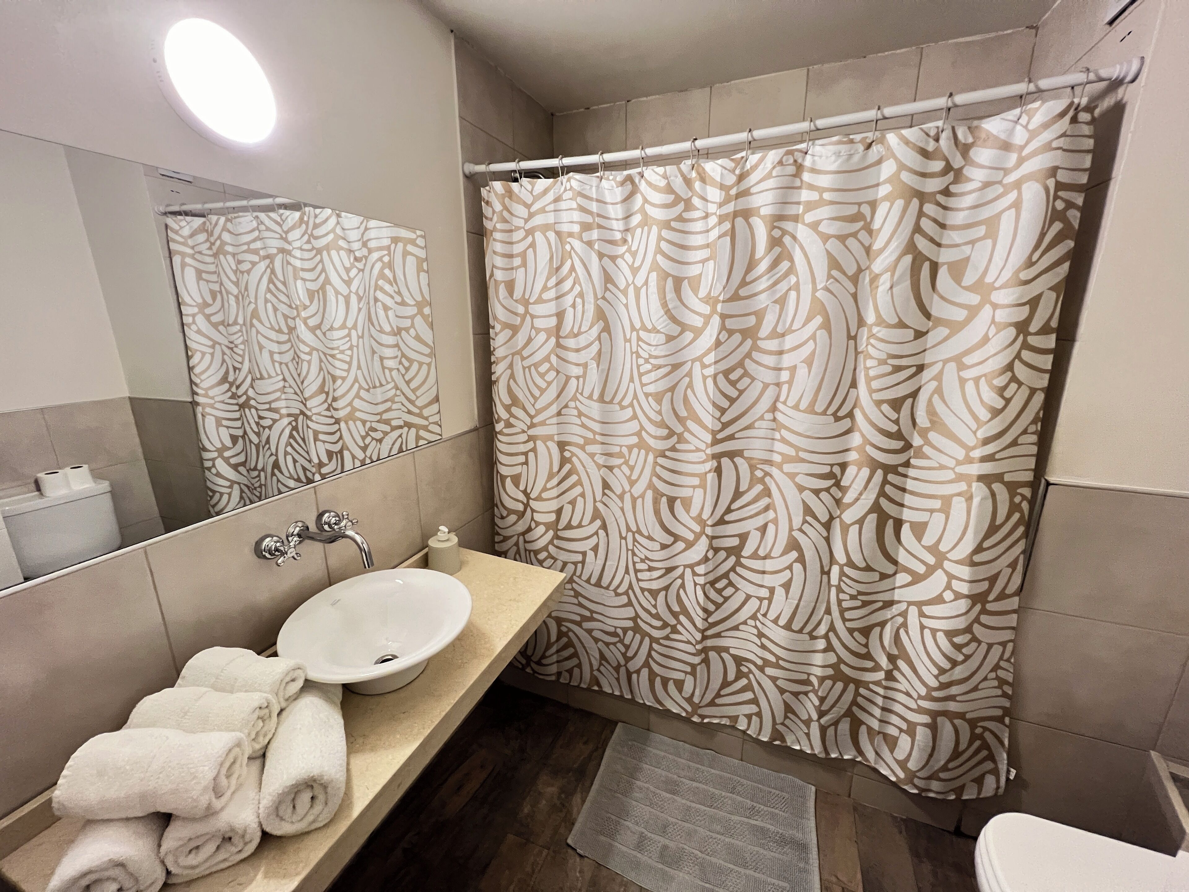 A bathroom with a white sink, a mirror, a towel rack with towels, a shower curtain with a leaf pattern, and a small rug on the floor.