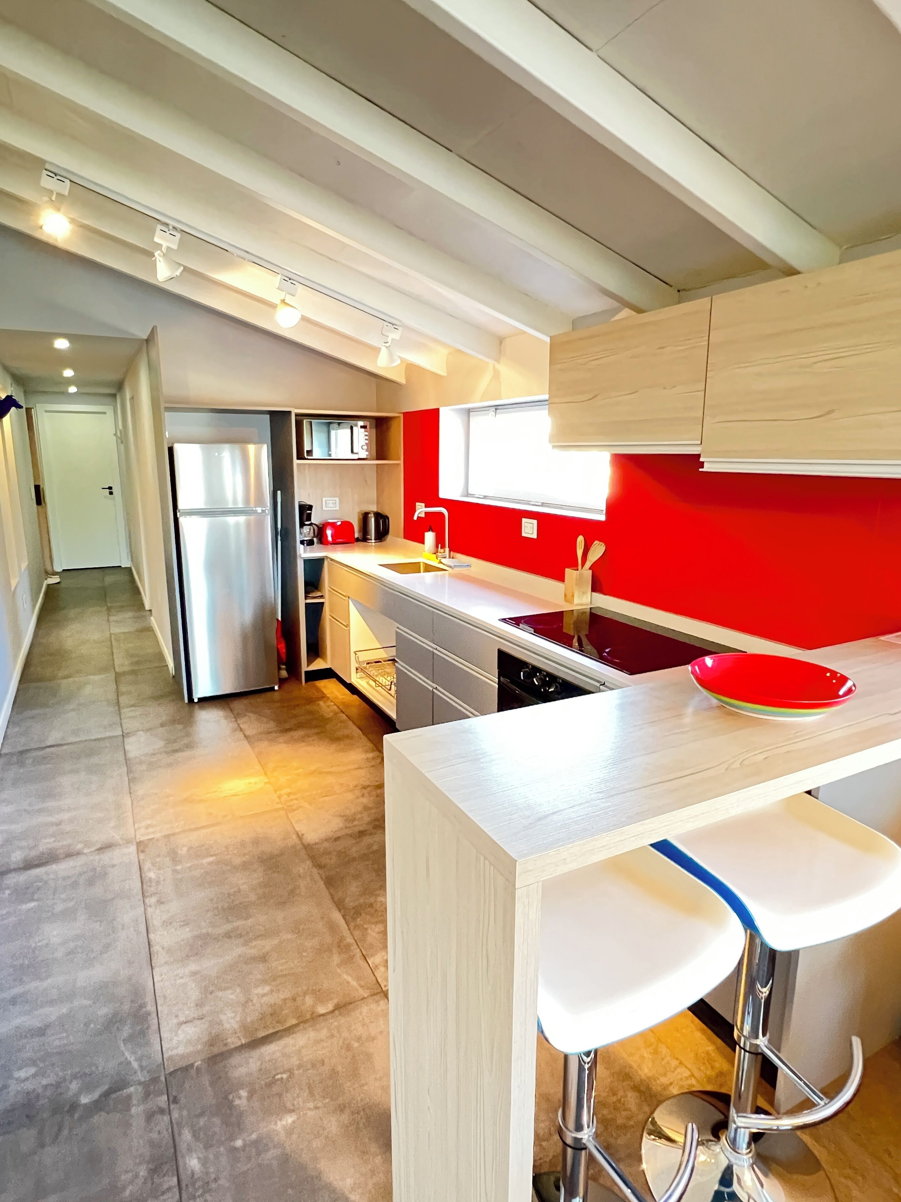 Una cocina moderna con un refrigerador de acero inoxidable, un microondas y un lavamanos. Hay una ventana con pared roja y una encimera de madera con un tazón rojo.