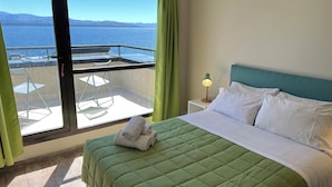 Une chambre d’hôtel avec un lit, une table de chevet, une lampe et une vue sur la mer grâce à de grandes fenêtres.