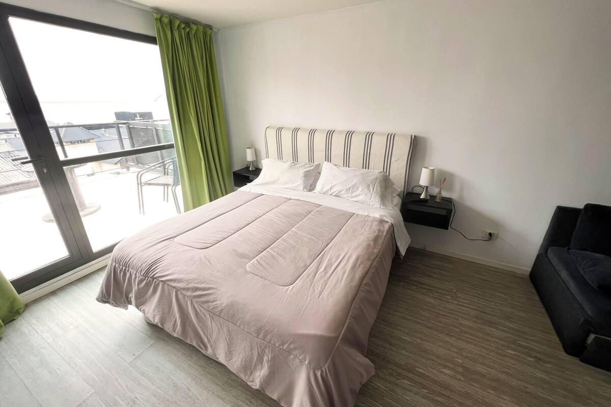 Una habitació d'hotel moderna amb un llit gran, una tauleta de nit amb una làmpada, un sofà negre i una vista a la ciutat a través d'una finestra gran.