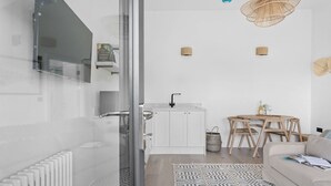 A modern, minimalist living space with a glass door leading to a bathroom, a small kitchen area with a sink and a round wooden table, and a beige sofa with a patterned rug. - The Sail Loft Suite - Sea View - Private patio (Brixham)