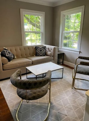Un salon avec un canapé beige, des fauteuils à motifs, une table basse blanche et de grandes fenêtres donnant sur un paysage verdoyant.