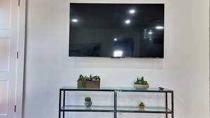 Ein Wohnzimmer mit einem an der Wand montierten Flachbildfernseher, einem Glastischchen mit Pflanzen und Holzböden.