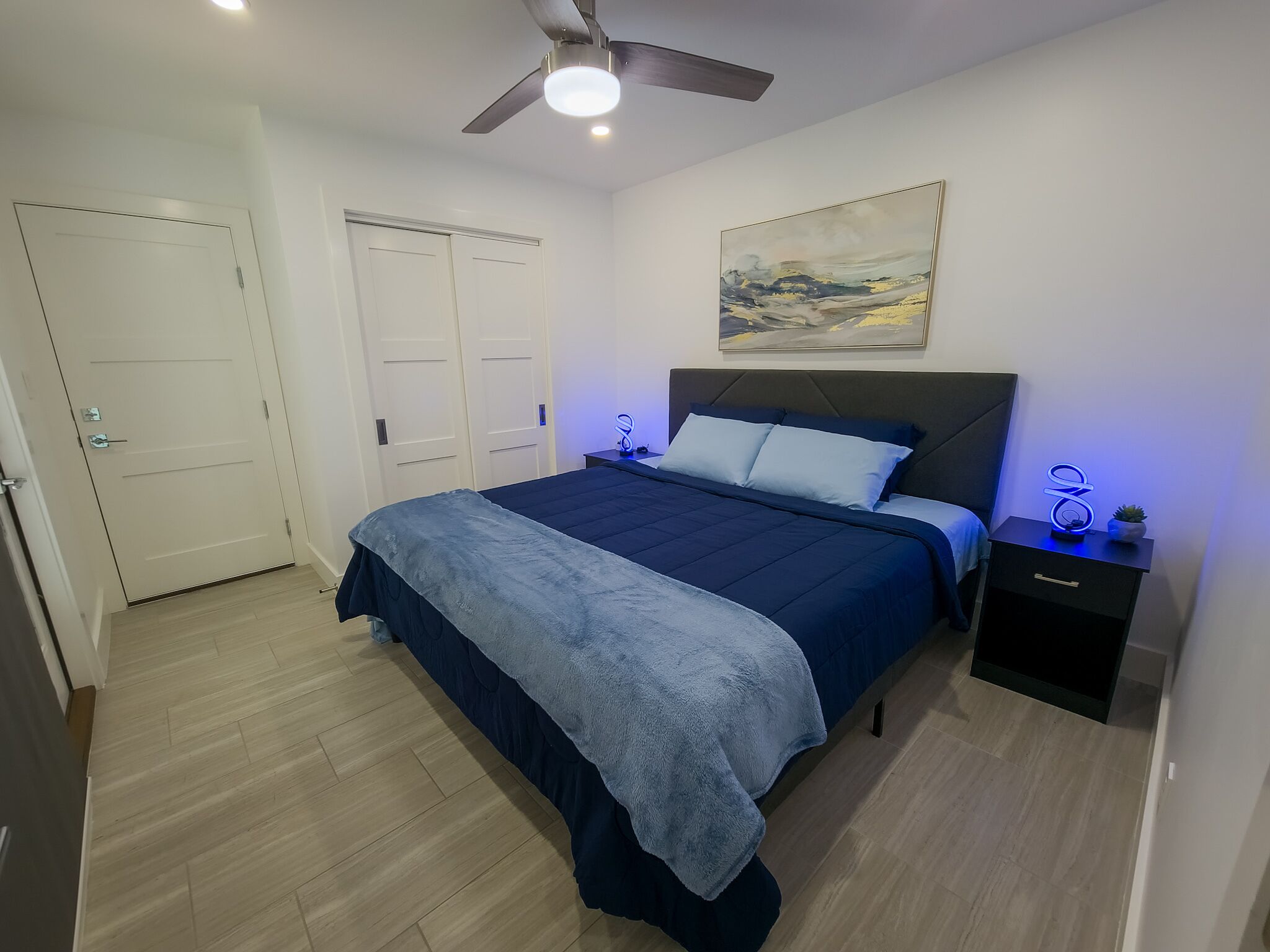 A bedroom with a bed, a nightstand, a ceiling fan, and a painting on the wall.