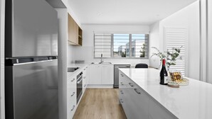 A modern kitchen with white cabinetry, a stainless steel refrigerator, a built-in dishwasher, and a large window providing natural light. - Azure Riverside DeluxeTownhouse (Sunshine Coast)