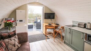 A cozy, well-lit room with a wooden ceiling, a dining area with wicker chairs, a small kitchen with a toaster and kettle, and a view of the outdoors through a door. - Cherry Pod 2 (Shrewsbury)