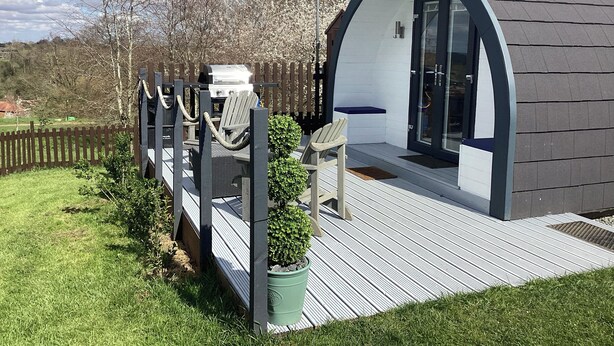 A small, modern, arched structure with a deck, a grill, and a chair. - Cherry Pod 2 (Shrewsbury)