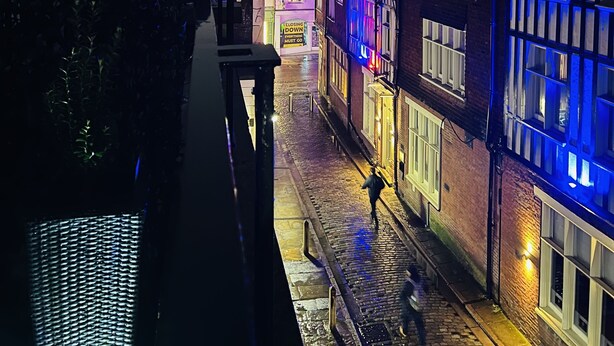 Uma rua estreita com edifícios em ambos os lados, alguns com janelas iluminadas.