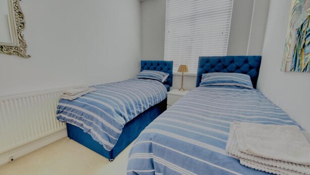 Quarto com duas camas, paredes brancas, radiador, janela com persianas e um quadro na parede.