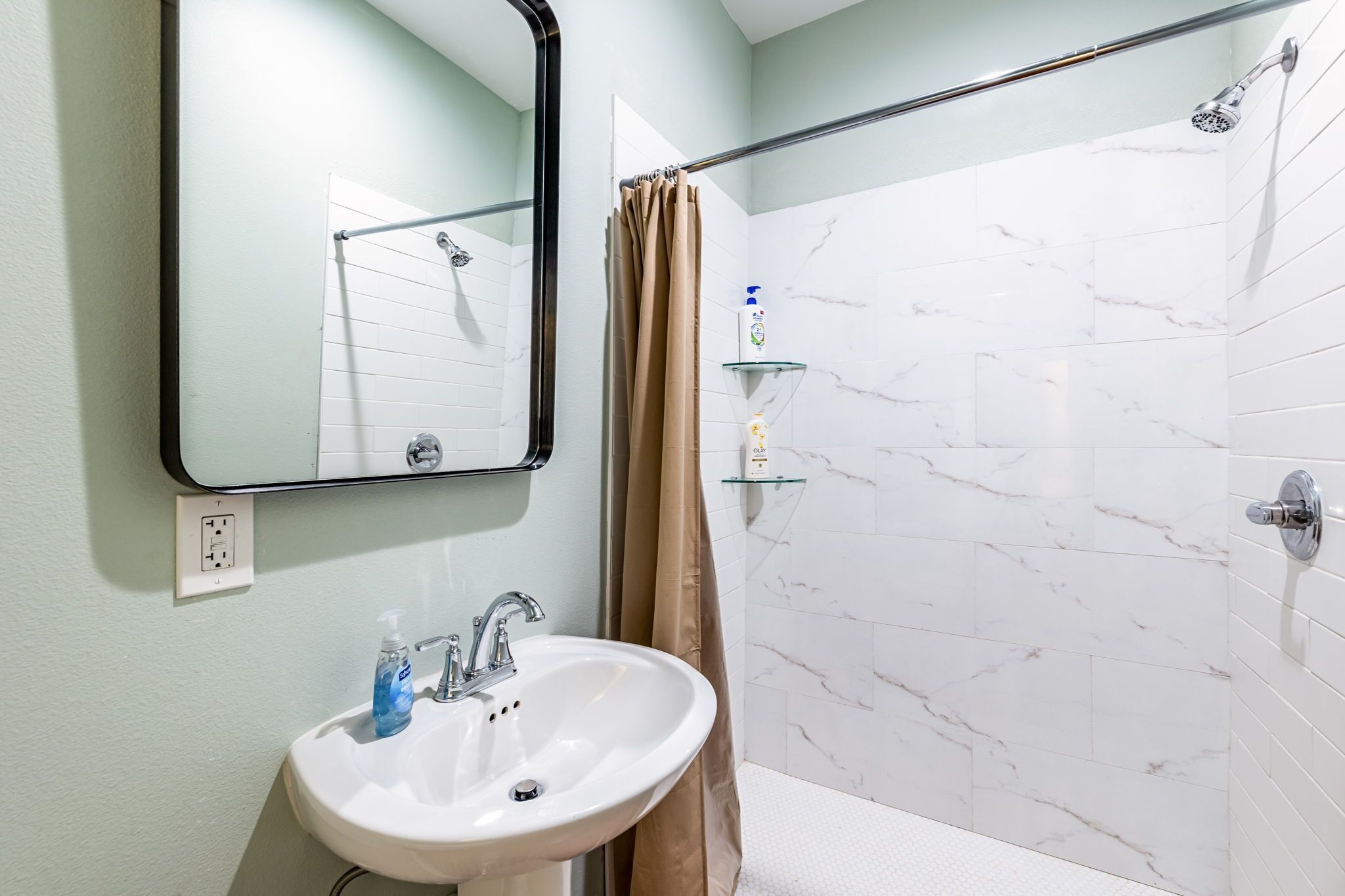 Un baño con un lavabo blanco, un espejo, una ducha con repisa de vidrio y una cortina color beige.