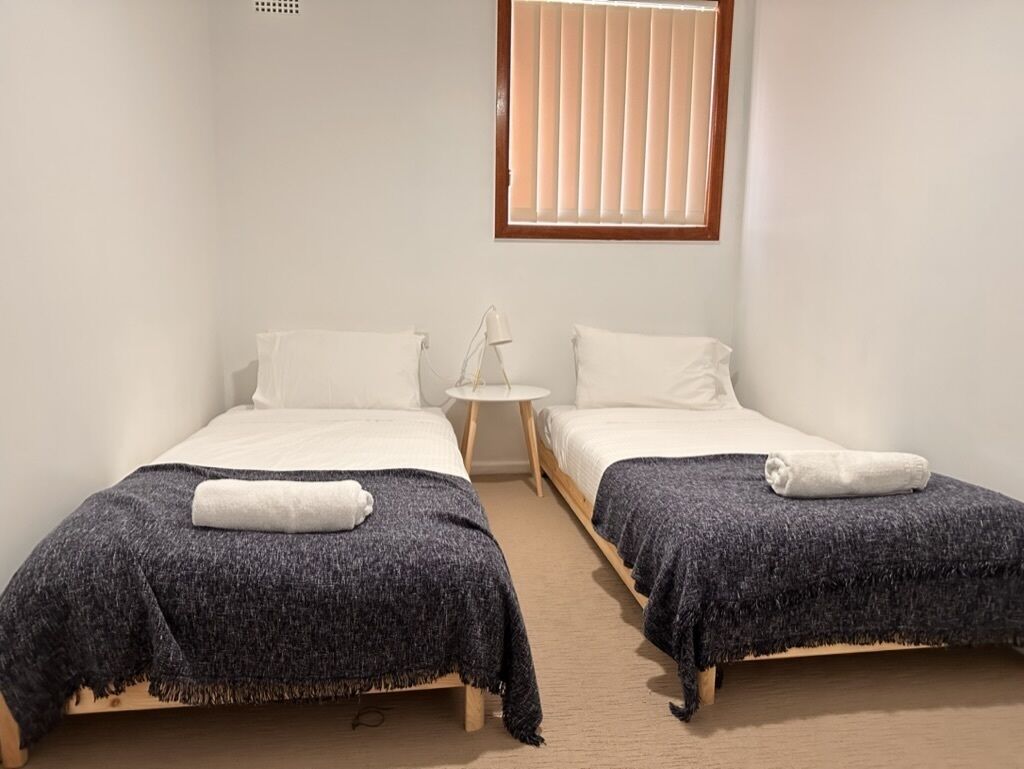 A room with two single beds, each with a white pillow and a dark grey blanket. There is a small round table between the beds with a lamp on it.
