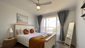 Un dormitorio con cama blanca, ventilador de techo, lámpara de noche, mesita redonda de mimbre, ventana con cortinas y un espejo.