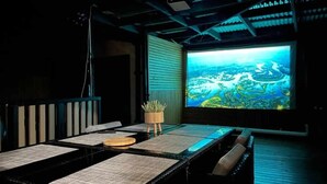 Una habitación oscura con una pantalla grande mostrando un paisaje pintoresco, una mesa con una planta y un proyector.