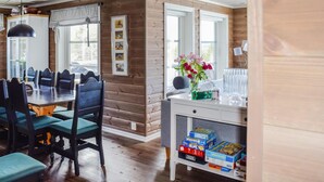 Interior acogedor de una cabaña con comedor que incluye sillas de madera y una mesa, una mesita auxiliar con libros y un jarrón de flores, y una ventana con vista a los árboles.