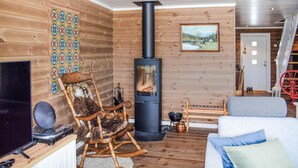 Una habitación acogedora con detalles de madera, una estufa de leña negra, un sillón mecedora, un sofá y un cuadro en la pared.