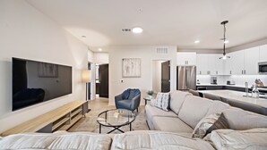 A modern living room with a large flat-screen TV, a sofa, a coffee table, and a kitchen area in the background. - Art house sawyer yards (Houston)