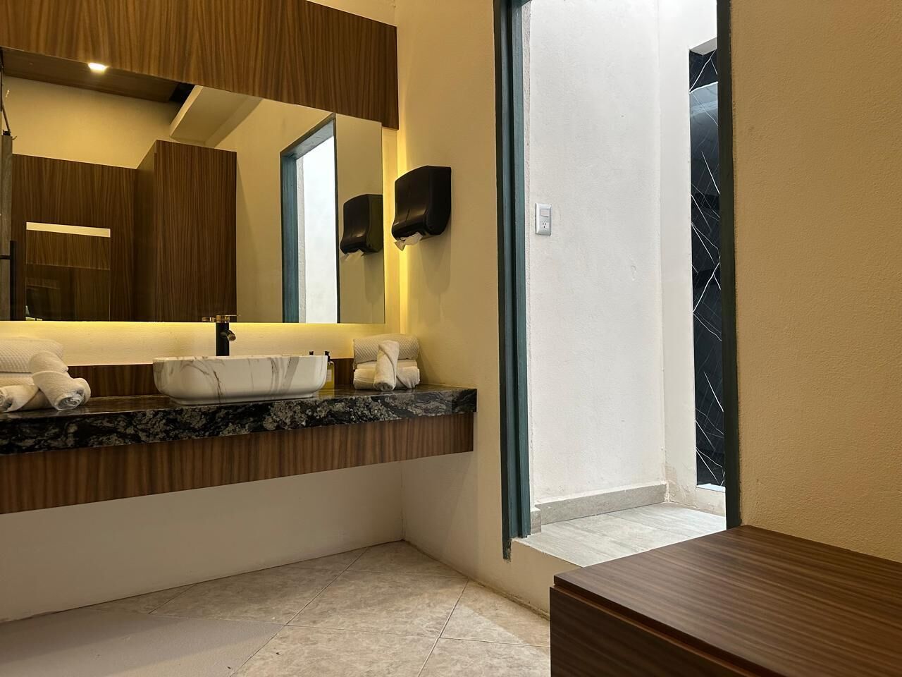 A modern bathroom with a marble countertop, a sink, and a large mirror. There are towels on the counter and a window with a view of the outside.