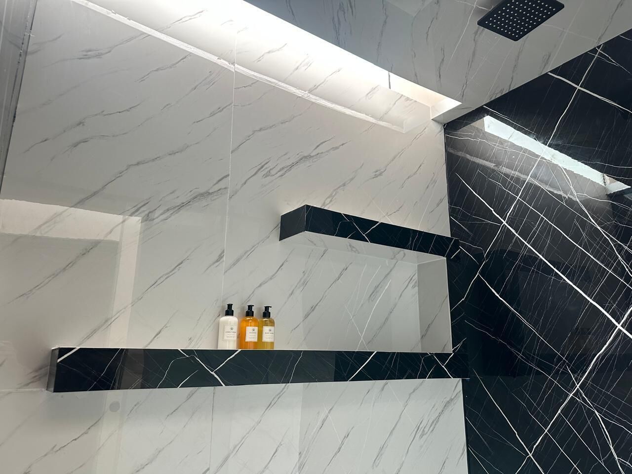 Un baño con paredes de mármol y una pared acentuada con azulejos negros. Hay dos botellas en un estante.