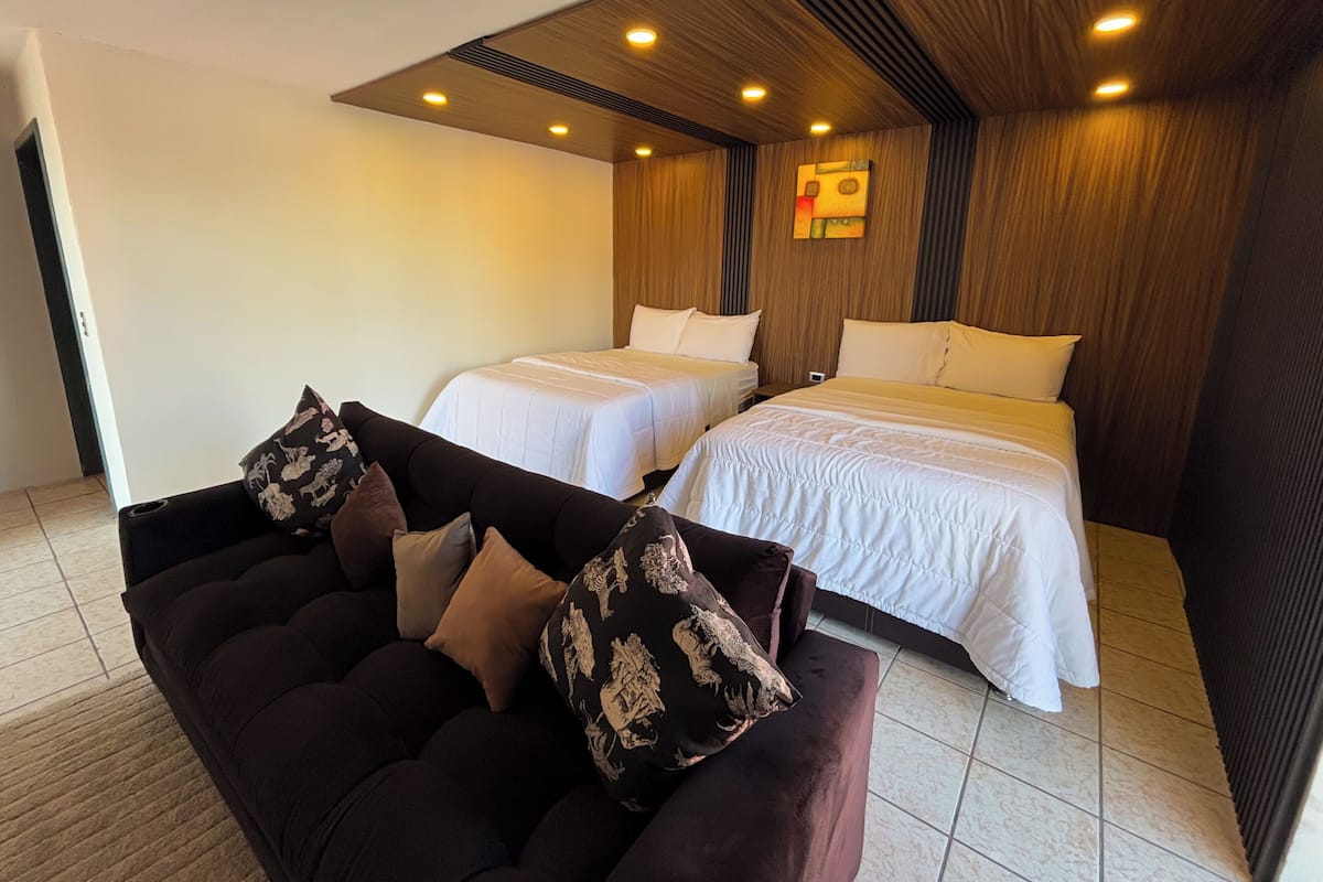 A hotel room with two beds, a sofa, and a wooden ceiling with recessed lighting.