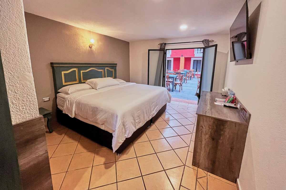 A hotel room with a bed, a small table, and a view of the outdoor seating area.