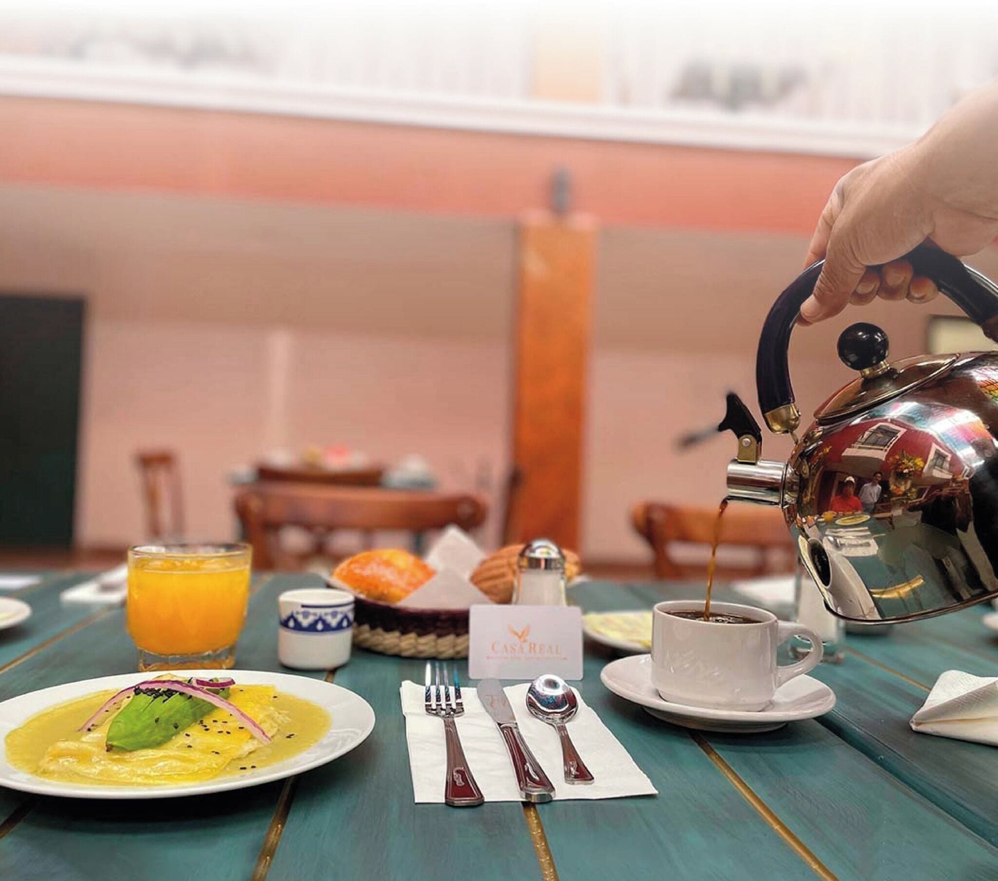 A breakfast setting with a teapot pouring coffee into a cup, a plate of food, a glass of orange juice, and a basket of bread.