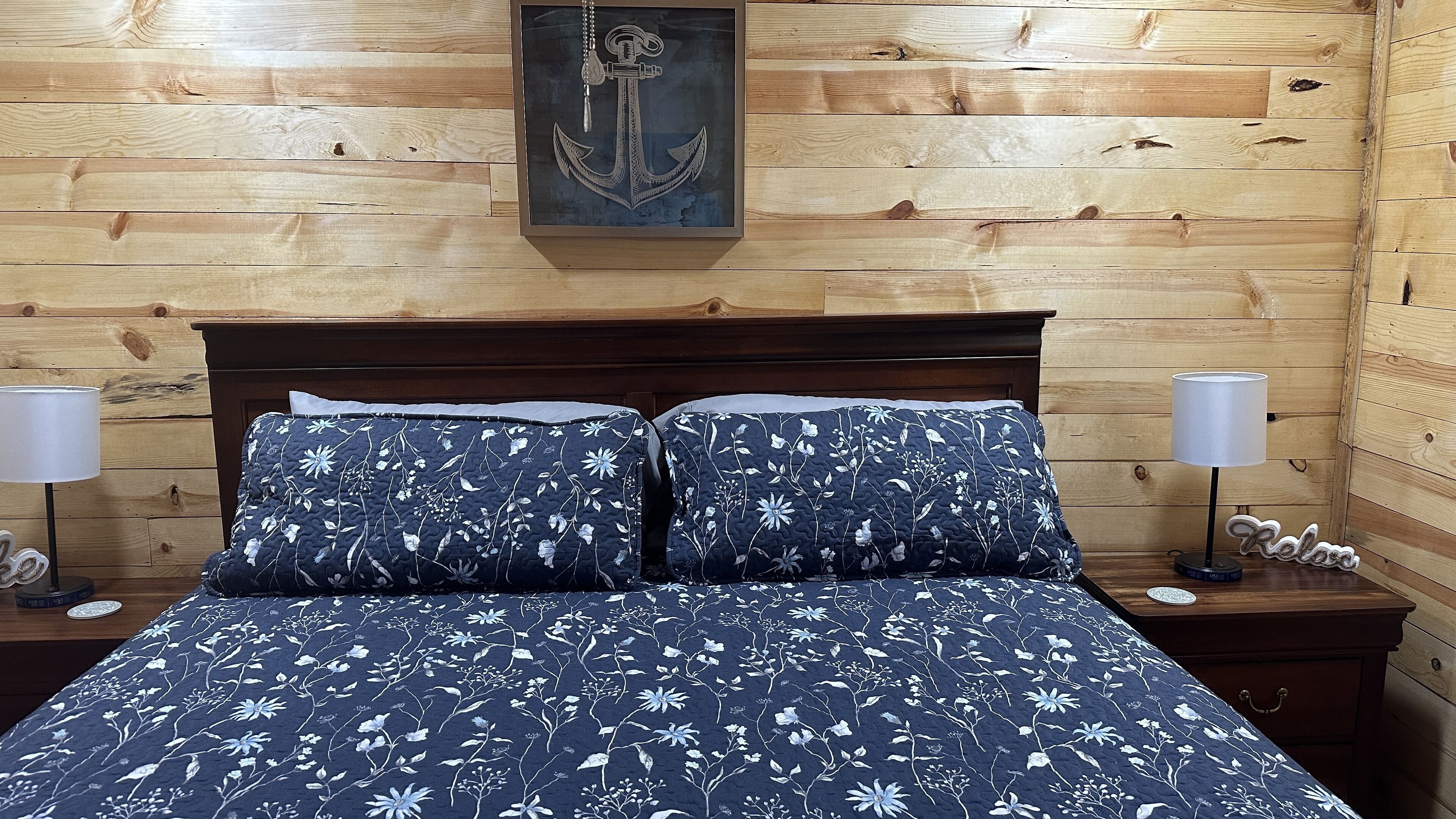 A wooden-paneled room with a bed, two bedside lamps, and a framed anchor artwork on the wall.