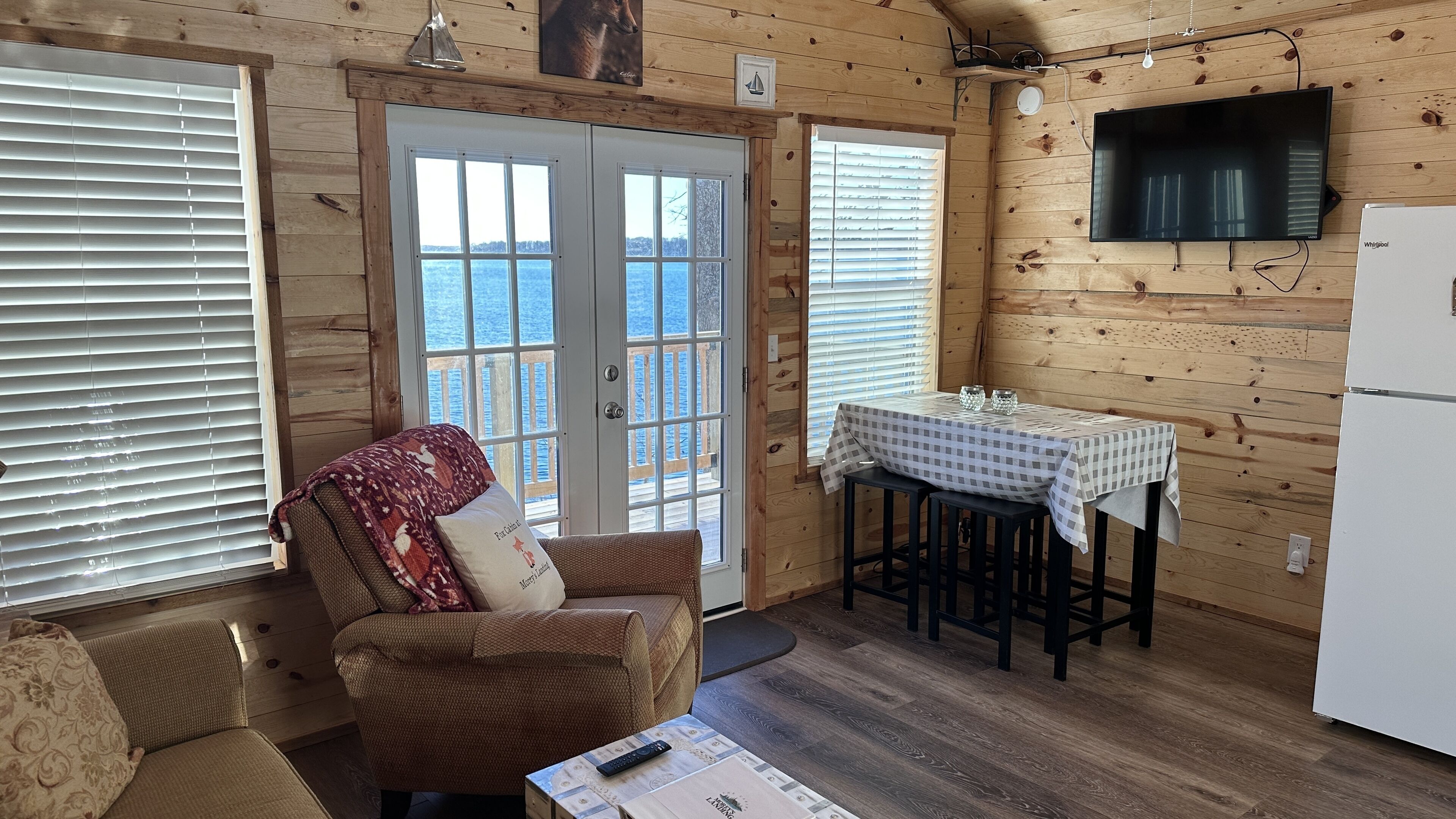 A cozy cabin interior with a wooden exterior, a dining area with a table and chairs, a sofa with a patterned throw, and a television mounted on the wall.