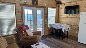 Interior acogedor de una cabaña con fachada de madera, una zona de comedor con mesa y sillas, un sofá con un plaid estampado, y un televisor montado en la pared.