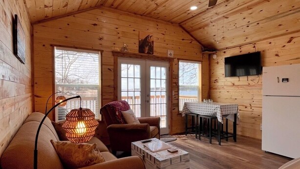 Interior acogedor de una cabaña con techo de madera, un sofá, una mesa de centro, una zona de comedor con mesa y sillas, y un televisor montado en la pared.