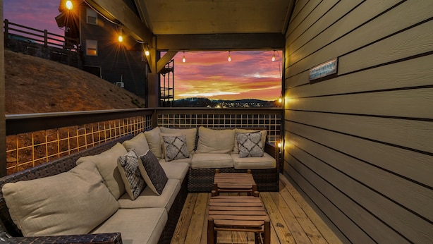 Un espace extérieur abrité avec des sièges, comprenant un canapé et un pouf, offrant une vue sur le coucher de soleil, et un panneau fixé au mur.