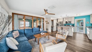A modern living room with a blue sectional sofa, a wooden coffee table, and a white armchair. There is a kitchen area with white cabinets and a blue accent wall.
