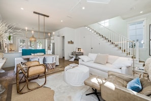 Amplio salón con sofá blanco, una mesa de centro y una silla de mimbre. Una escalera con barandillas blancas conduce a otro nivel.