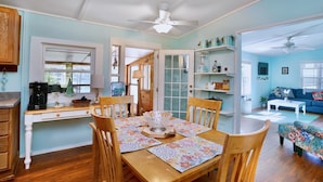 A dining area with a wooden table, chairs, and a ceiling fan. There is a kitchen area with cabinets and a coffee maker.