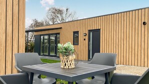 A modern outdoor patio with a table and chairs, a decorative plant, and a wooden building with large glass doors in the background. - 2 bedroom stunning home in Heinkenszand (Heinkenszand)