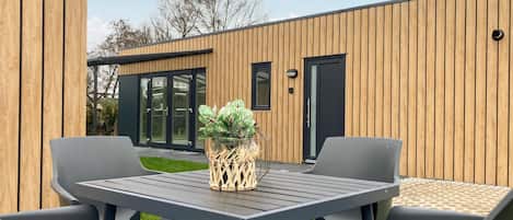 A modern outdoor patio with a table and chairs, a decorative plant, and a wooden building with large glass doors in the background.