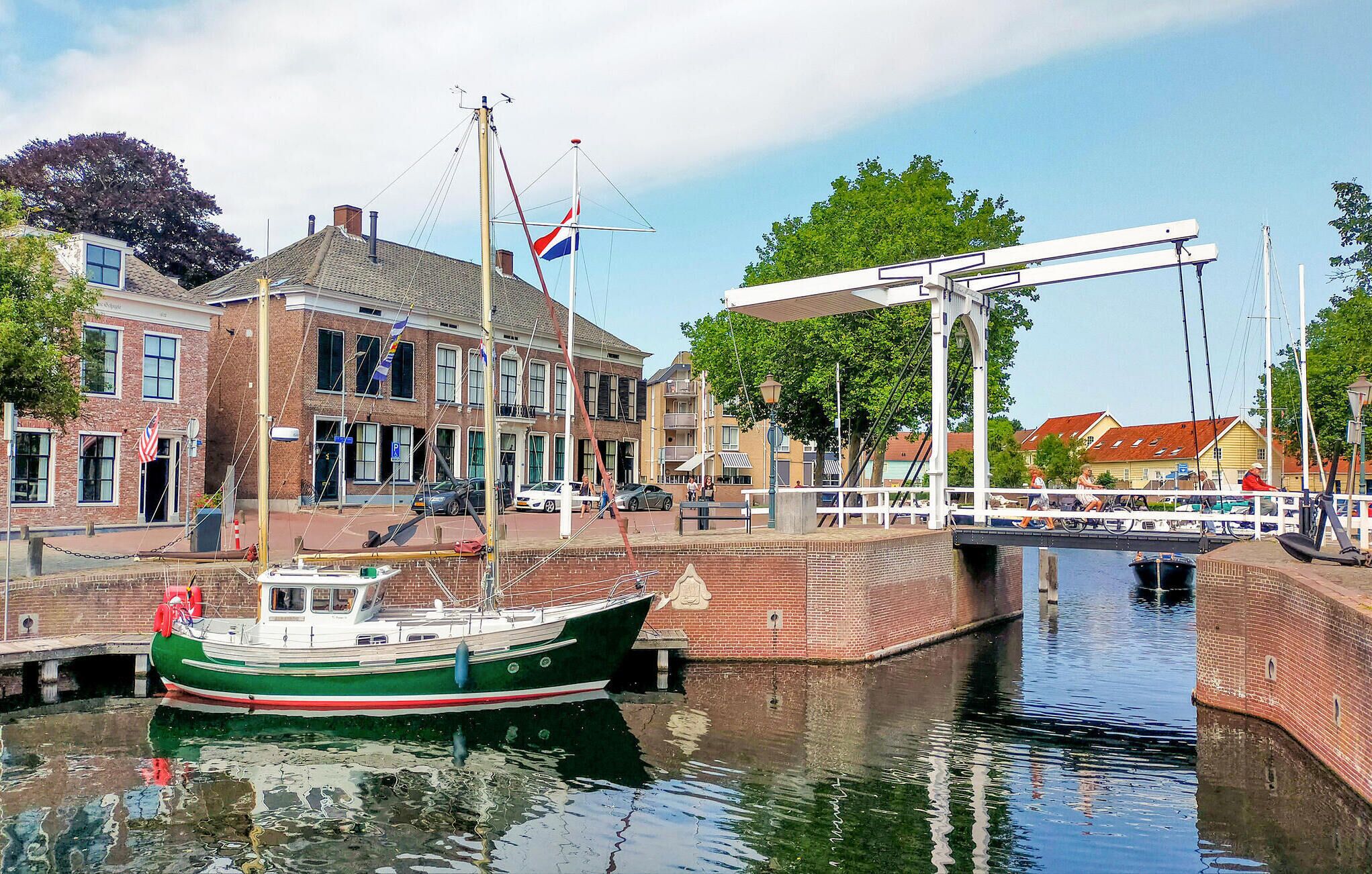 Een gracht met een groene boot afgemeerd, een brug en gebouwen met vlaggen.