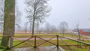 Une terrasse en bois avec une rambarde en métal, d’où l’on peut admirer un paysage brumeux parsemé d’arbres et où l’on aperçoit, au loin, un bâtiment rouge.
