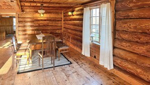 L’intérieur d’un chalet rustique en rondins de bois, avec une table et des chaises en bois, une fenêtre avec des rideaux et un luminaire au plafond.