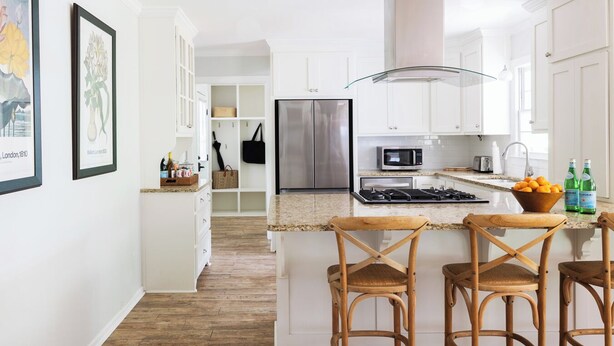 A modern kitchen with white cabinets, a stainless steel refrigerator, a microwave, and a gas stove. There are wicker bar stools and a fruit bowl on the counter. - Game Day Retreat minutes to Downtown and Sanford Stadium (Athens)