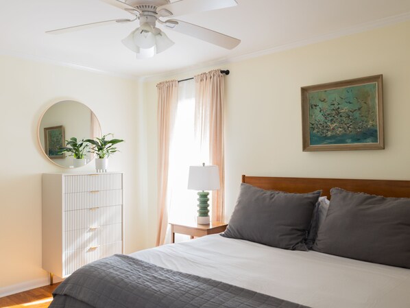 Une chambre à coucher avec un lit, un ventilateur de plafond, une commode, une lampe, une fenêtre avec des rideaux et un tableau au mur.
