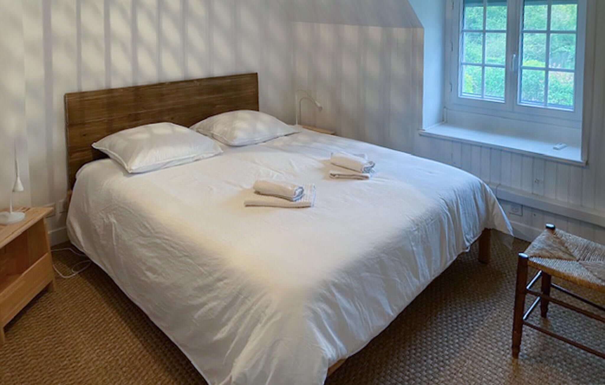 Un lit bien fait, avec du linge de lit et des serviettes blanches, une tête de lit en bois, une table de chevet avec une lampe et une fenêtre donnant sur un paysage verdoyant.