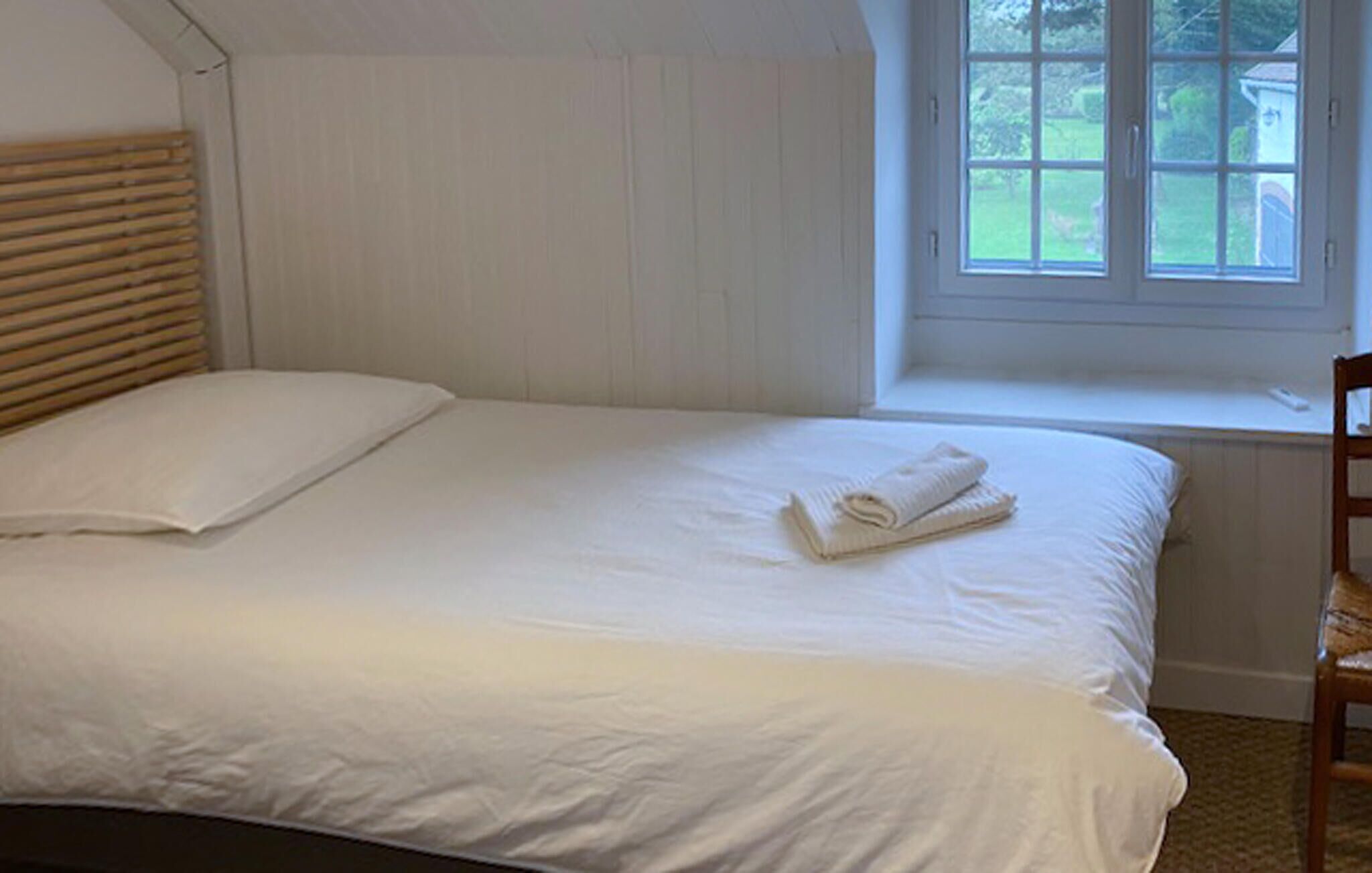 Un lit bien fait, avec du linge de lit blanc et une serviette pliée, une tête de lit en bois et une fenêtre donnant sur un paysage verdoyant.