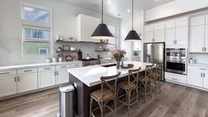 Una cucina moderna con mobili bianchi, un'isola centrale con sgabelli da bar, elettrodomestici in acciaio inossidabile e illuminazione a sospensione.