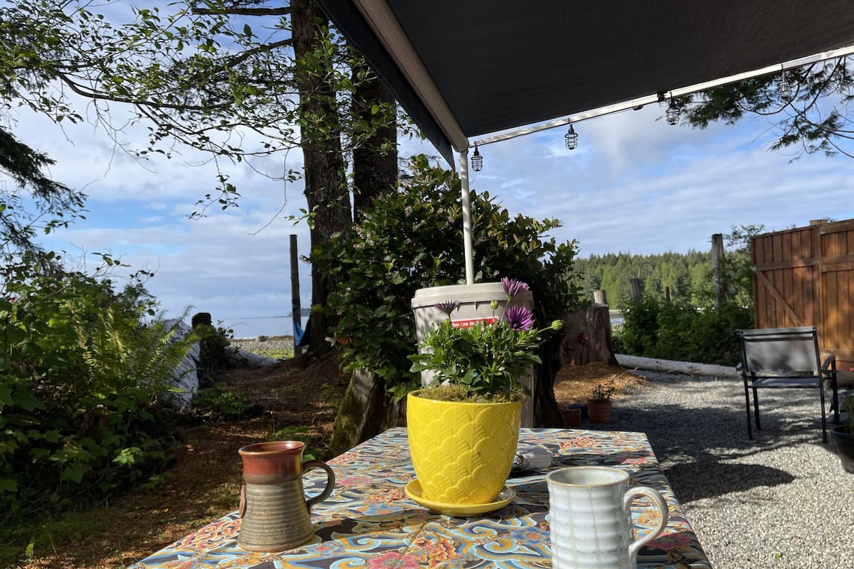 Un área de estar al aire libre con mantel estampado, una maceta amarilla con flores, un jarro de cobre y una taza blanca.