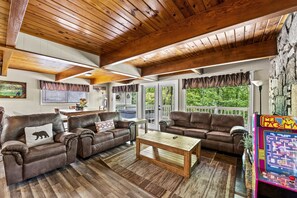 A cozy living room with wooden ceiling beams, leather sofas, a wooden coffee table, and large windows with curtains.