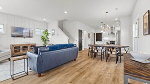 Amplia sala de estar con un sofá gris, una mesa de comedor de madera y una escalera que lleva a una cocina.