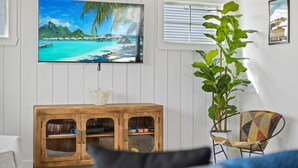 Una sala de estar con un mueble de madera, un televisor que muestra una playa tropical, una planta en maceta y un sillón estampado.