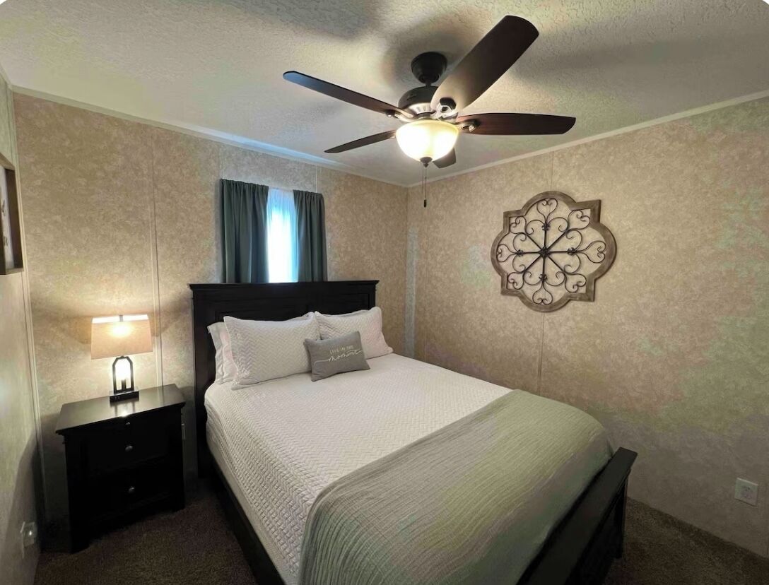 Une chambre à coucher comprenant un lit, une table de chevet, un élément de décoration murale et un ventilateur de plafond.