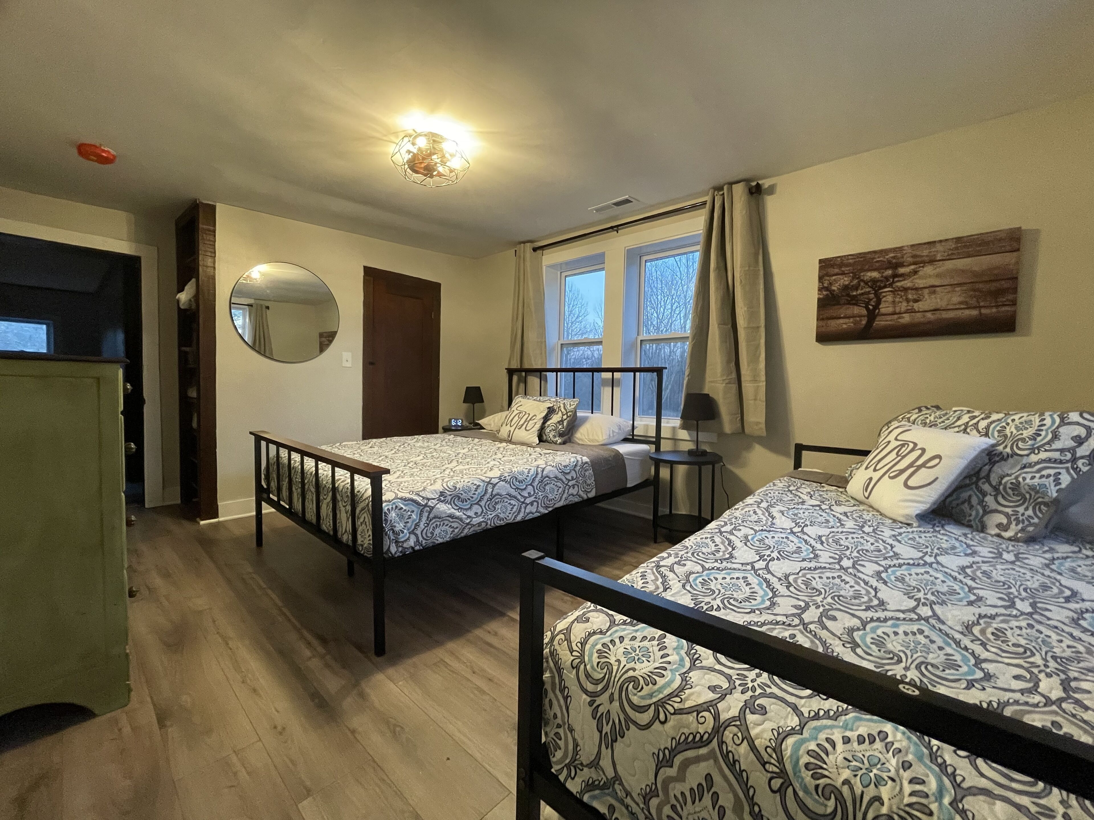 A bedroom with two beds, a bedside table with a lamp, a mirror, and a window with curtains.