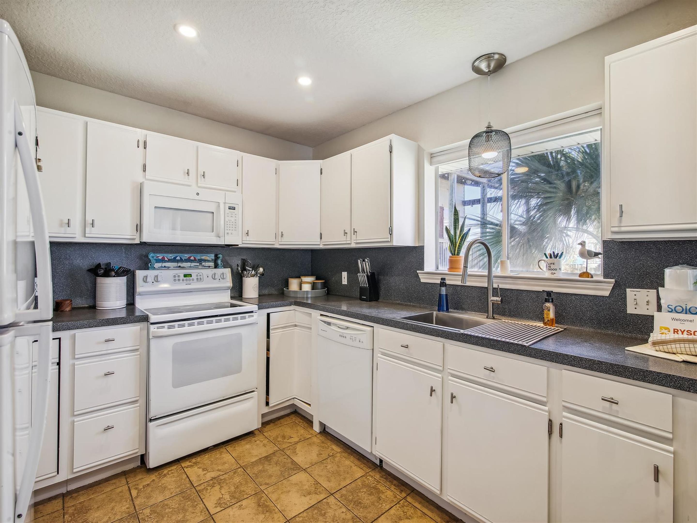 A modern kitchen with white cabinets, a stainless steel sink, and a window with a view of palm trees.